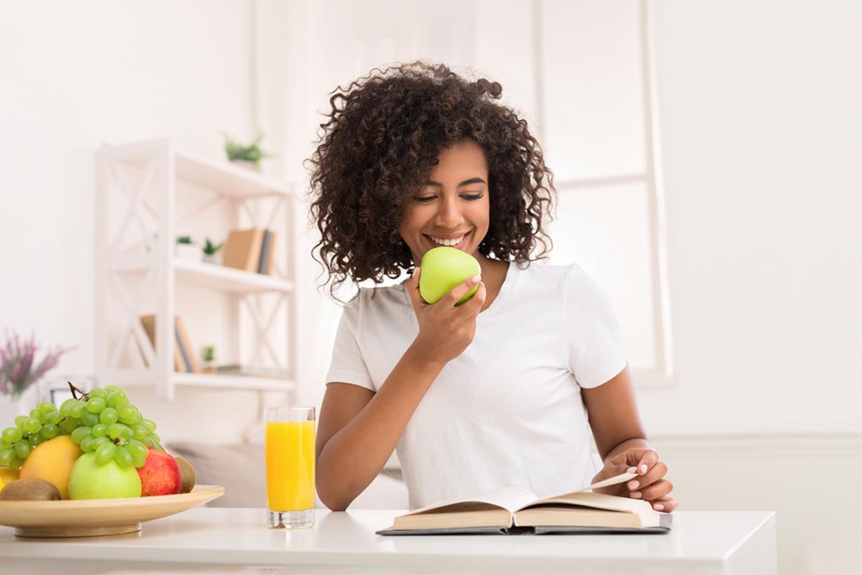 A woman sitting at a desk with one hand holding an apple while the other hand holds a page of a book.