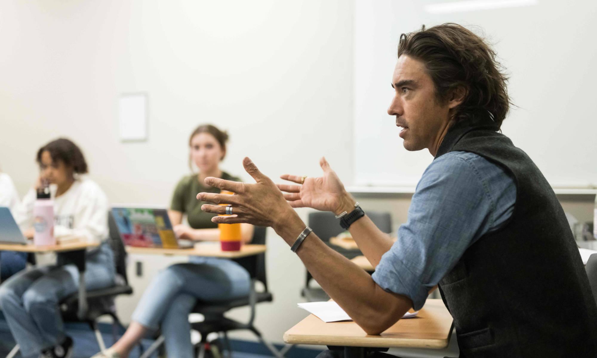 Jack Downey seated at a desk in a classroom teaching with two students seated with their laptops out of focus in the background.