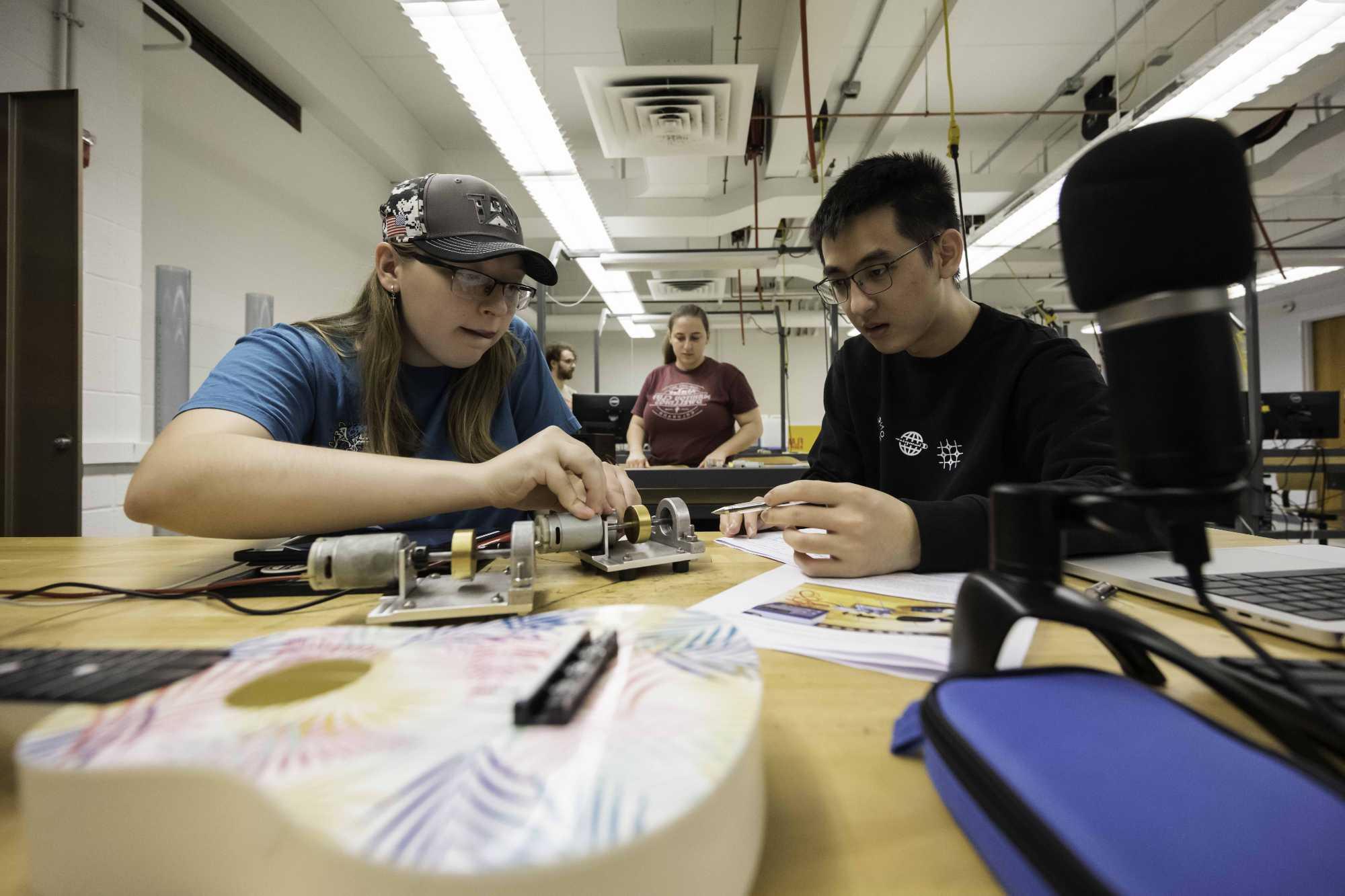 Two students lean over a mechanical 工程师ing lab bench with various instruments and tools in the foreground and two other students in the background.