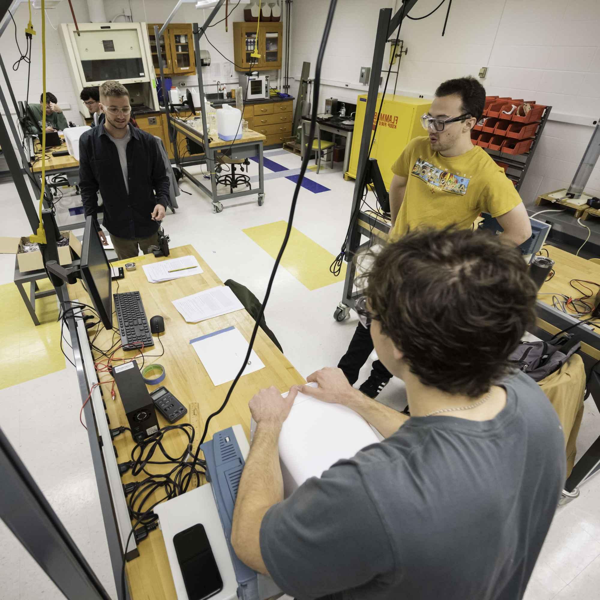 Three mechanical 工程师ing students stand around a table and conduct experiments during a repeatability and statistics lab.