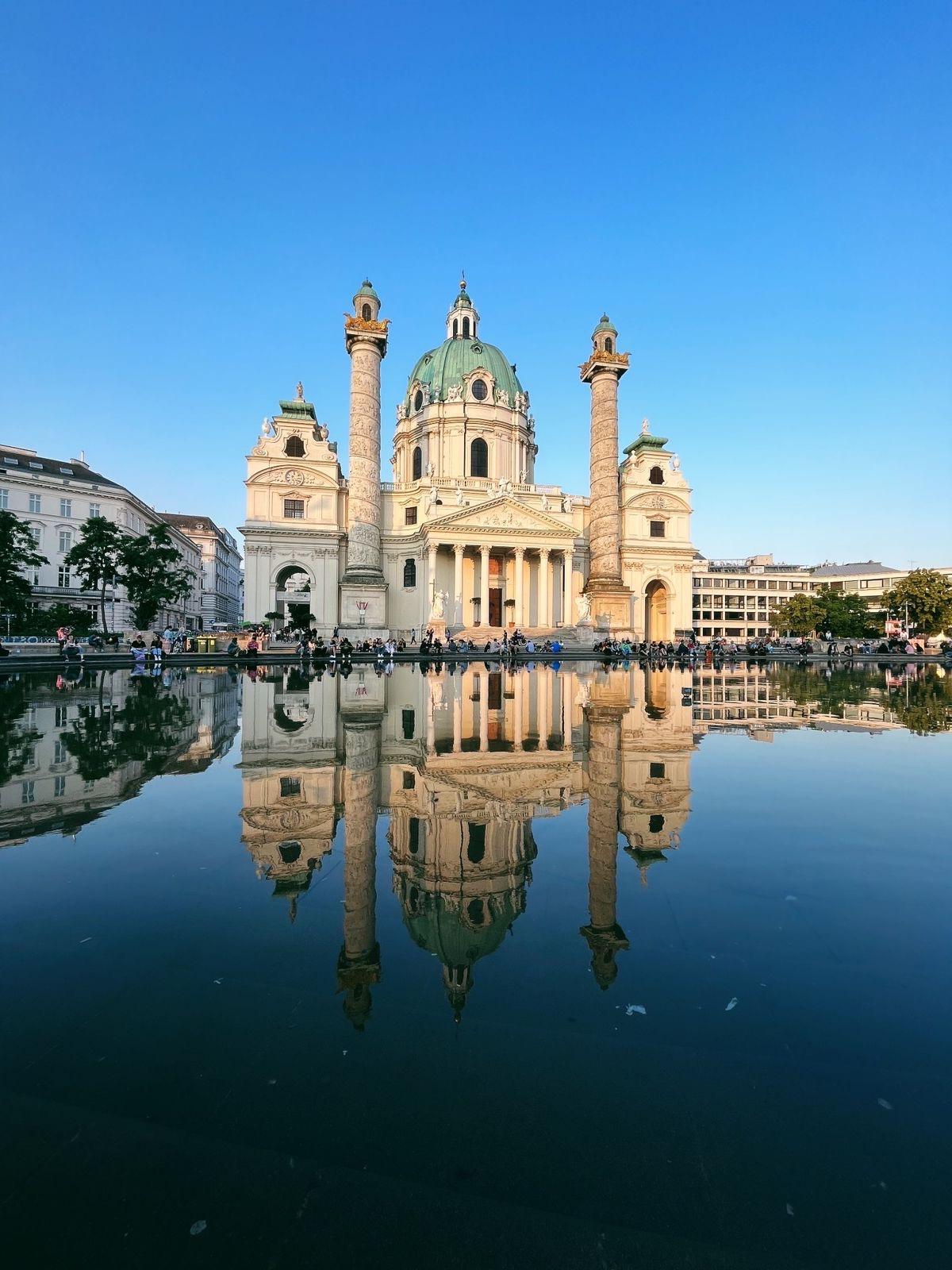 A grand baroque-style white cathedral with a large green dome and twin columns stands against a clear blue sky. 这座建筑在宁静中映出美丽的倒影, 前景是一池清澈的水, 人们聚集在它的基地周围.