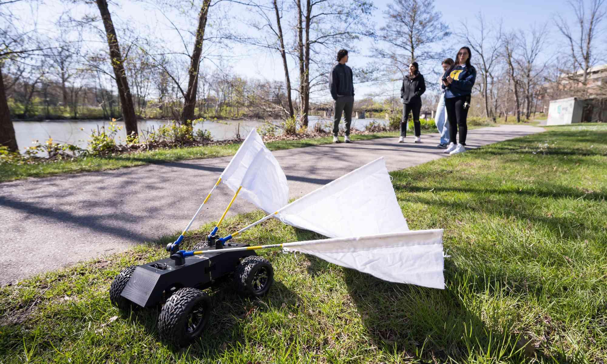 A radio controlled car with white flannel flags attached and four 学生 in the background controlling it on a bike path.