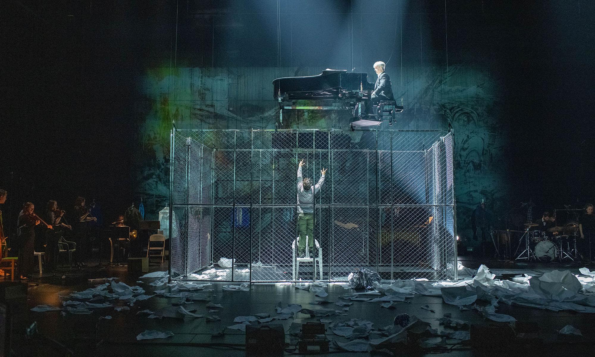 Man in cage reaches up toward piano player in scene from Paper Pianos.