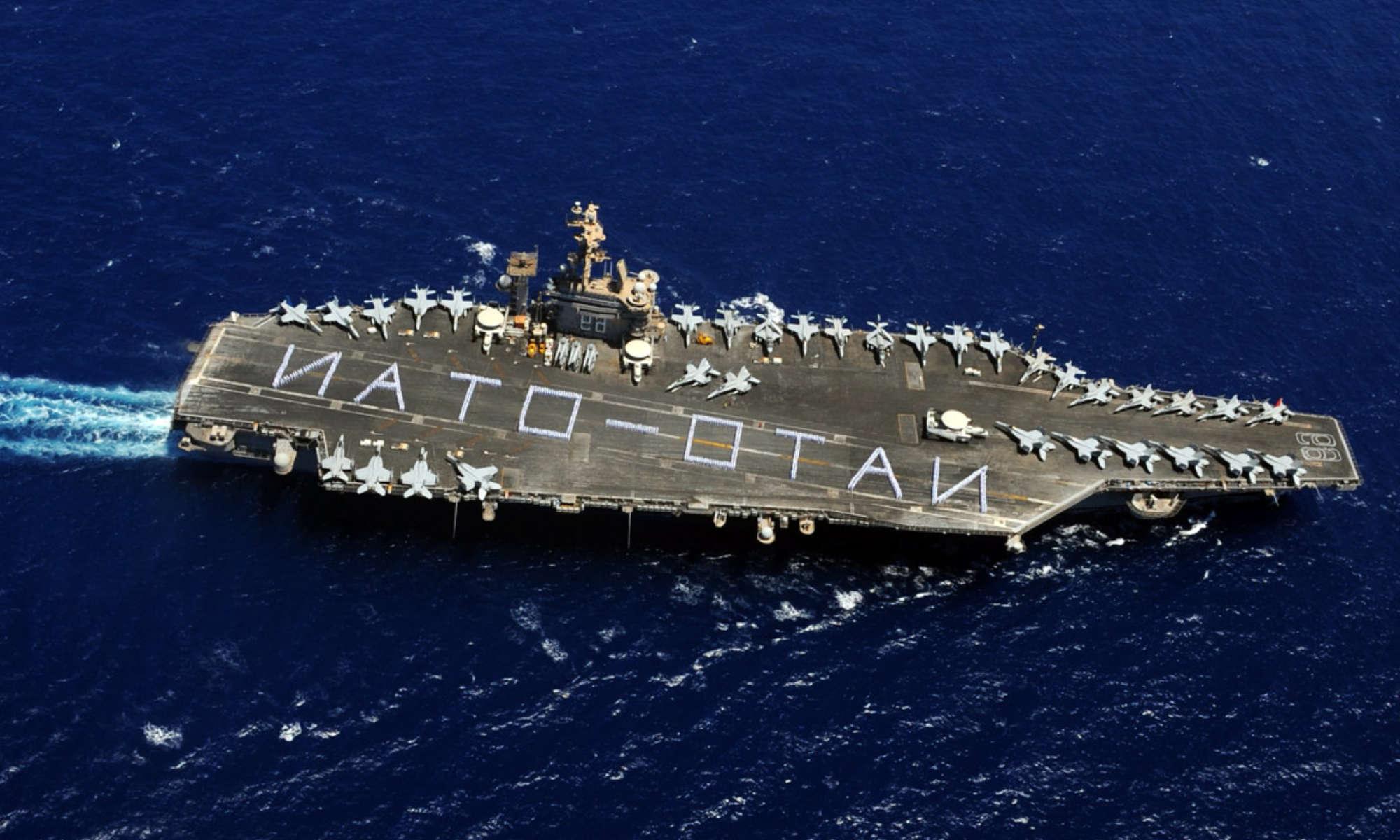 Aerial view of an aircraft carrier at sea with sailors spelling out the words NATO and OTAN.