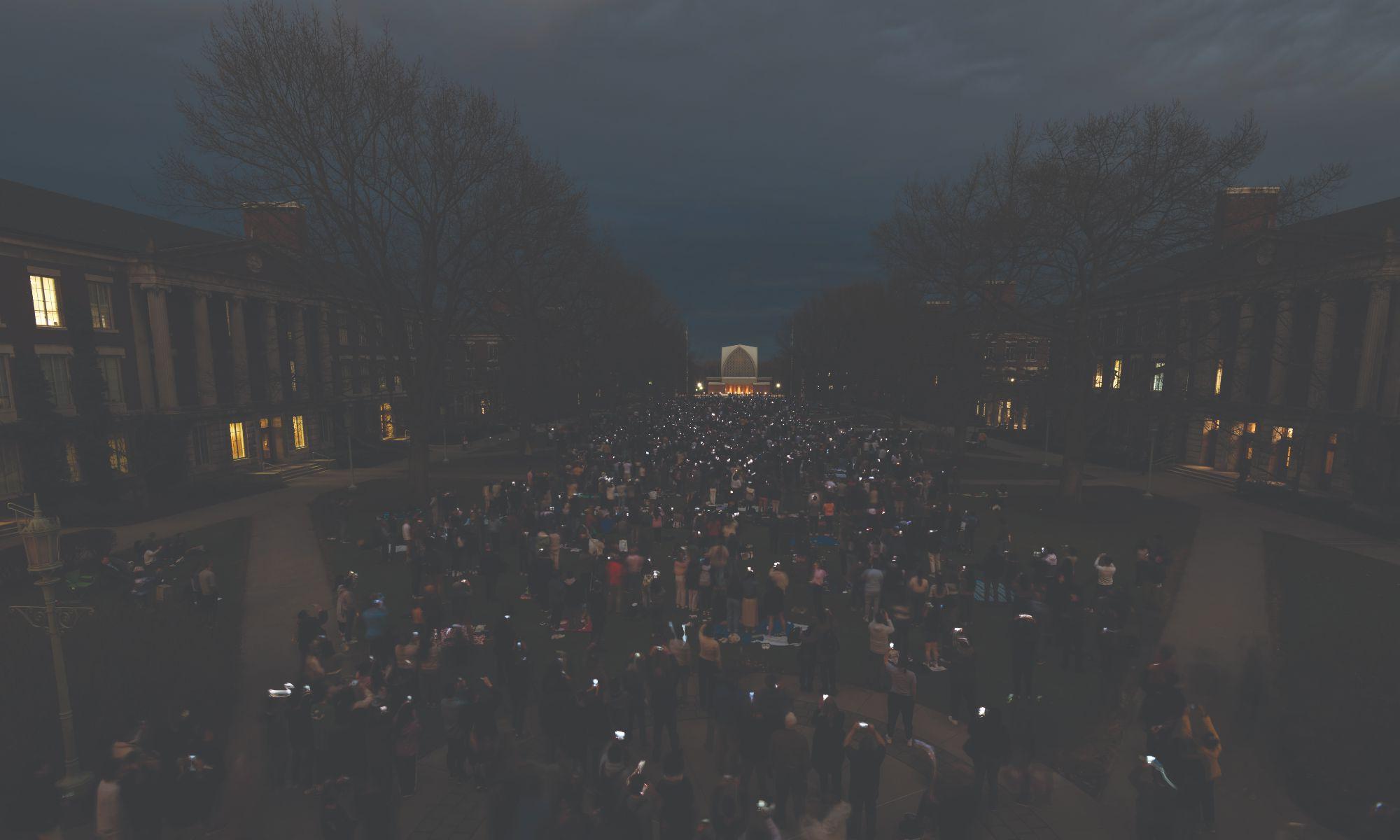 A large crowd of people gathers in an outdoor courtyard at dusk, holding up lit candles or lights. Trees surround the area and buildings with lit windows line both sides. The sky is darkening, and the scene appears peaceful and solemn.