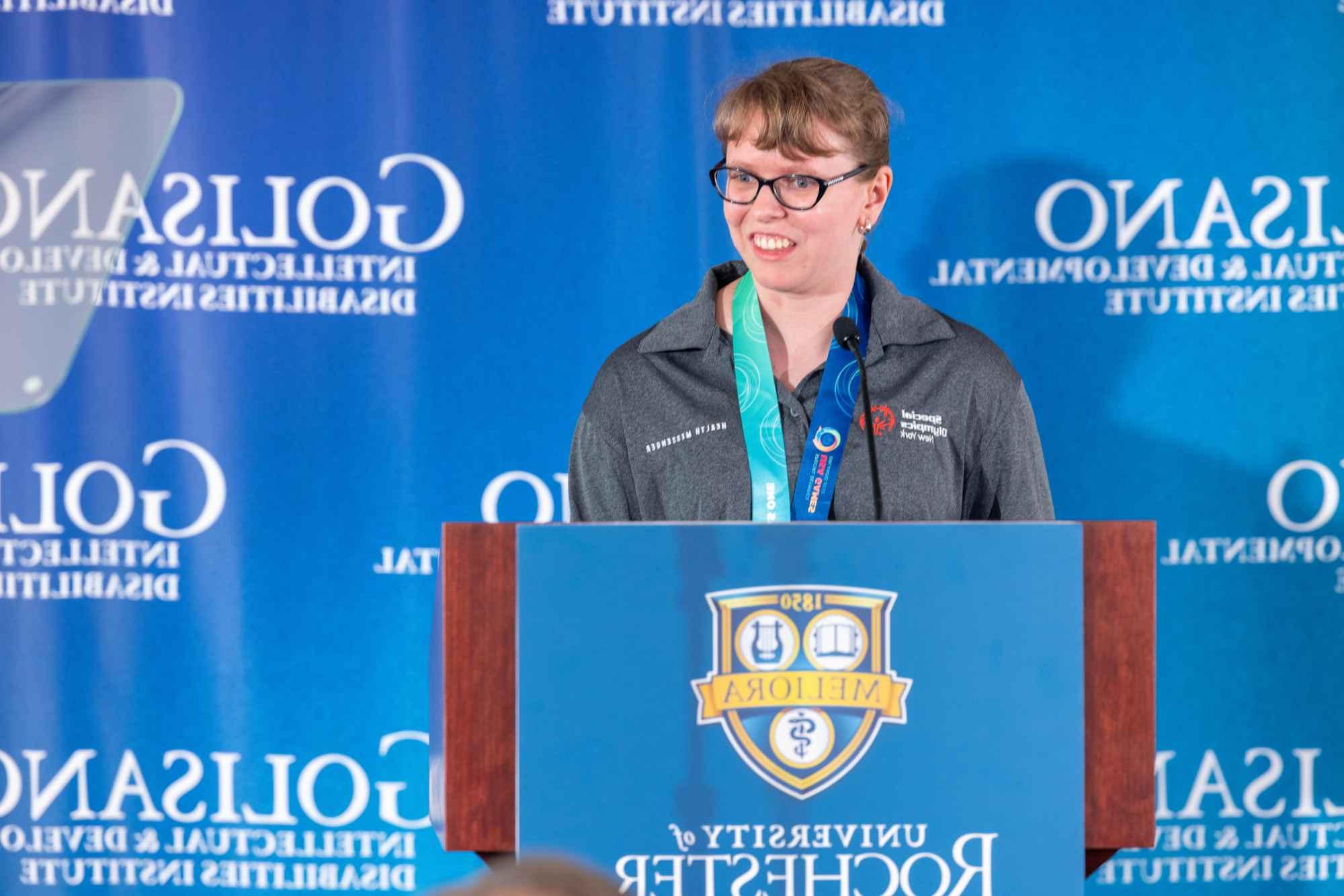 A female Special Olympics gold medalist and ambassador stands at a 澳门威尼斯人网上赌场 podium in front of a blue backdrop for the Golisano Intellectual and Developmental Disabilities Institute. 