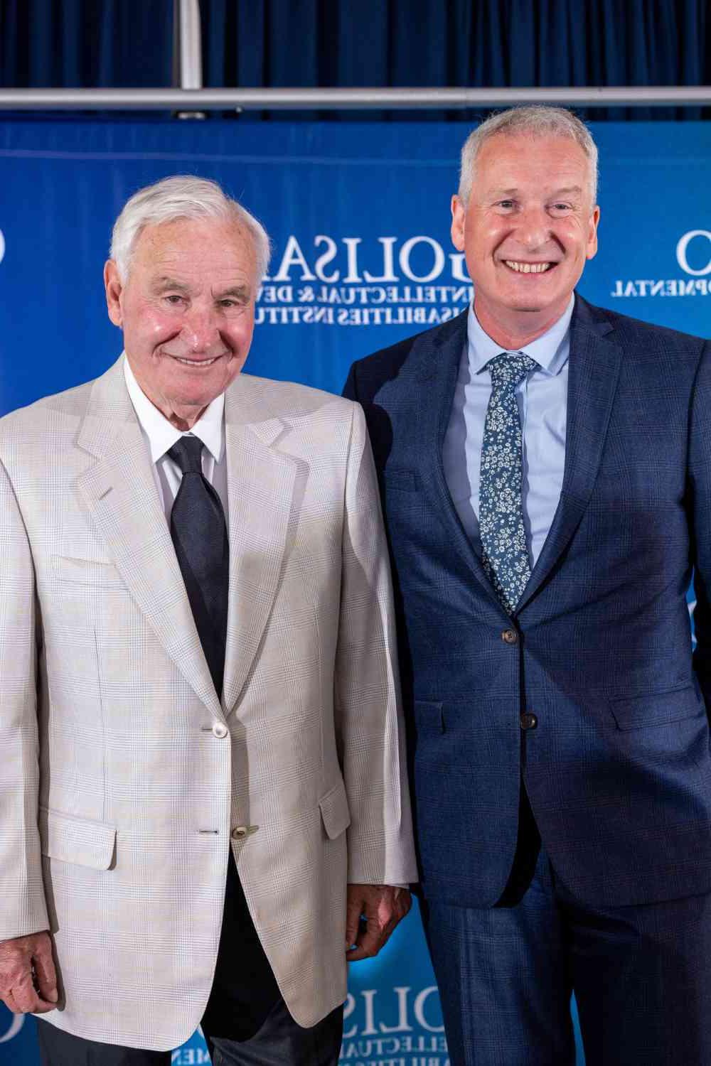 约翰·福克斯著 and 汤姆Golisano in front of a blue backdrop featuring the 澳门威尼斯人网上赌场 logo for the Golisano Intellectual and Developmental Disabilities Institute. 