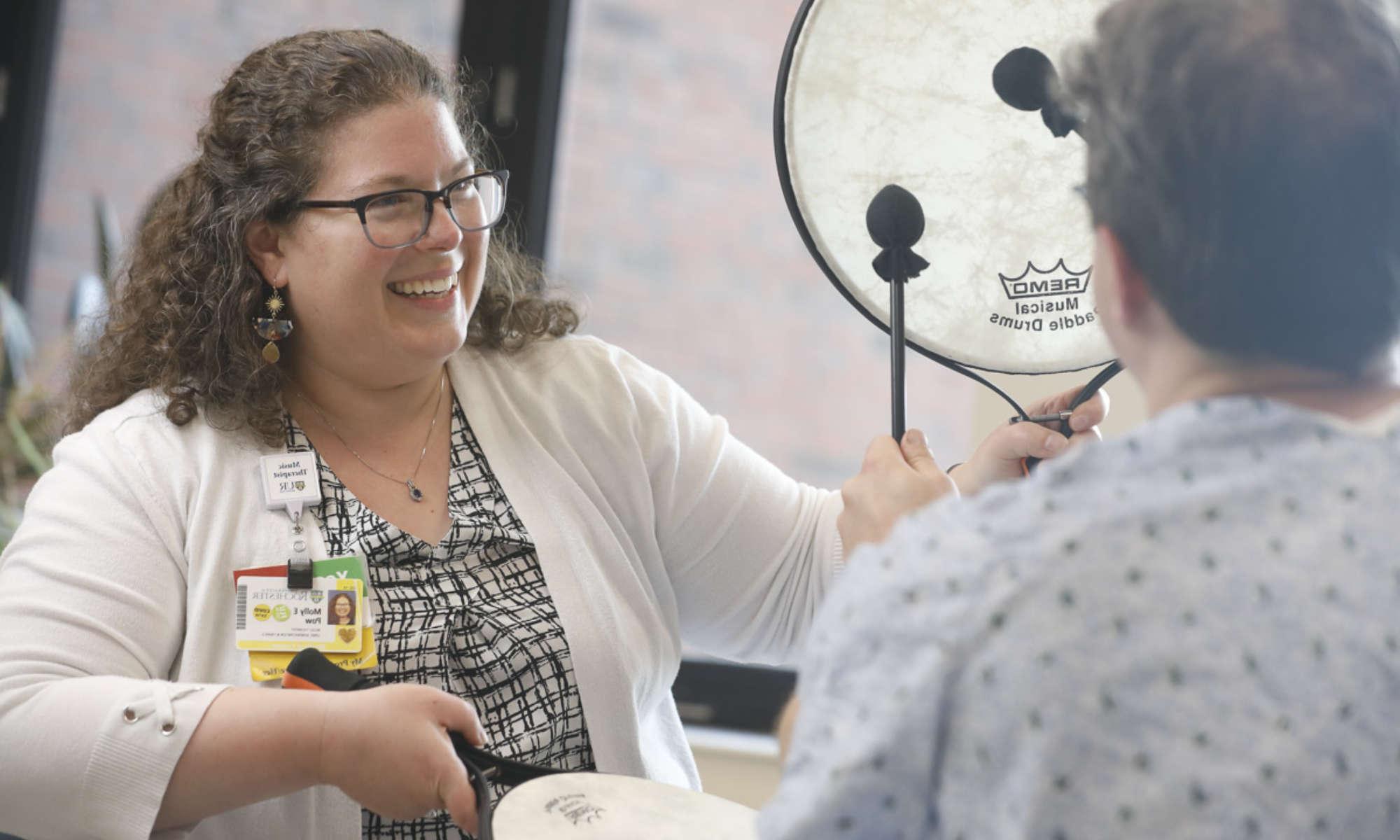 A music therapist smiles and holds paddle drums while a patient in a hospital gown seen from behind uses paddle sticks to beat the drums.