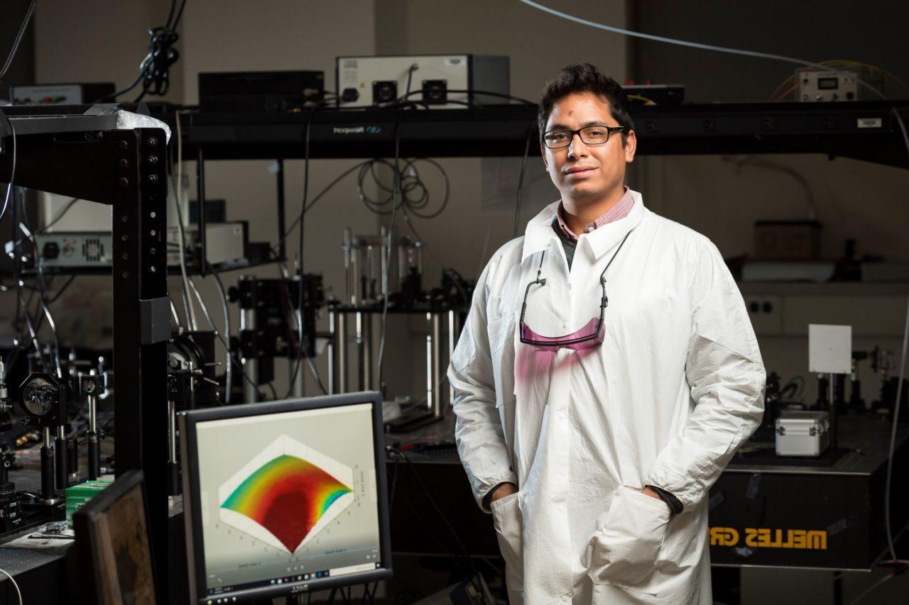 A University of Rochester researcher in a lab, wearing a lab coat and goggles, in front of a data visualization chart on a computer